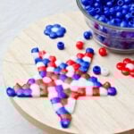 Red white and blue melted beads in the shape of a star sitting on a circular piece of wood next to a bowl of blue beads.