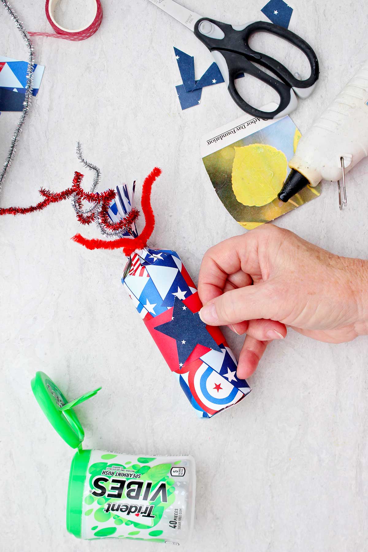 Person placing a blue patterned piece of scrapbook paper in the shape of a star on a finished 4th of July treat box.