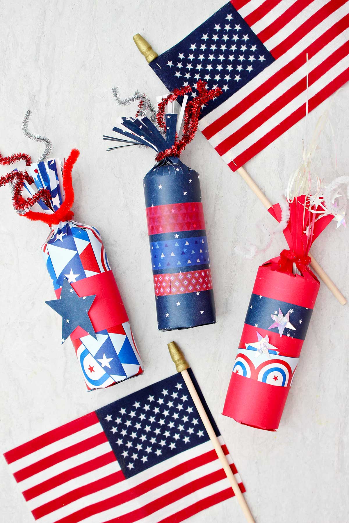 Three 4th of July firecracker treat boxes decorated with red white and blue paper and washi tape with two small American flags as decoration.