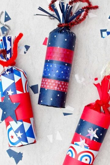 Three 4th of July firecracker treat boxes decorated with red white and blue paper and washi tape with some scraps of festive paper scattered around.