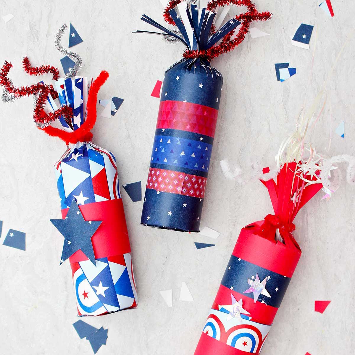 Three 4th of July firecracker treat boxes decorated with red white and blue paper and washi tape with some scraps of festive paper scattered around.