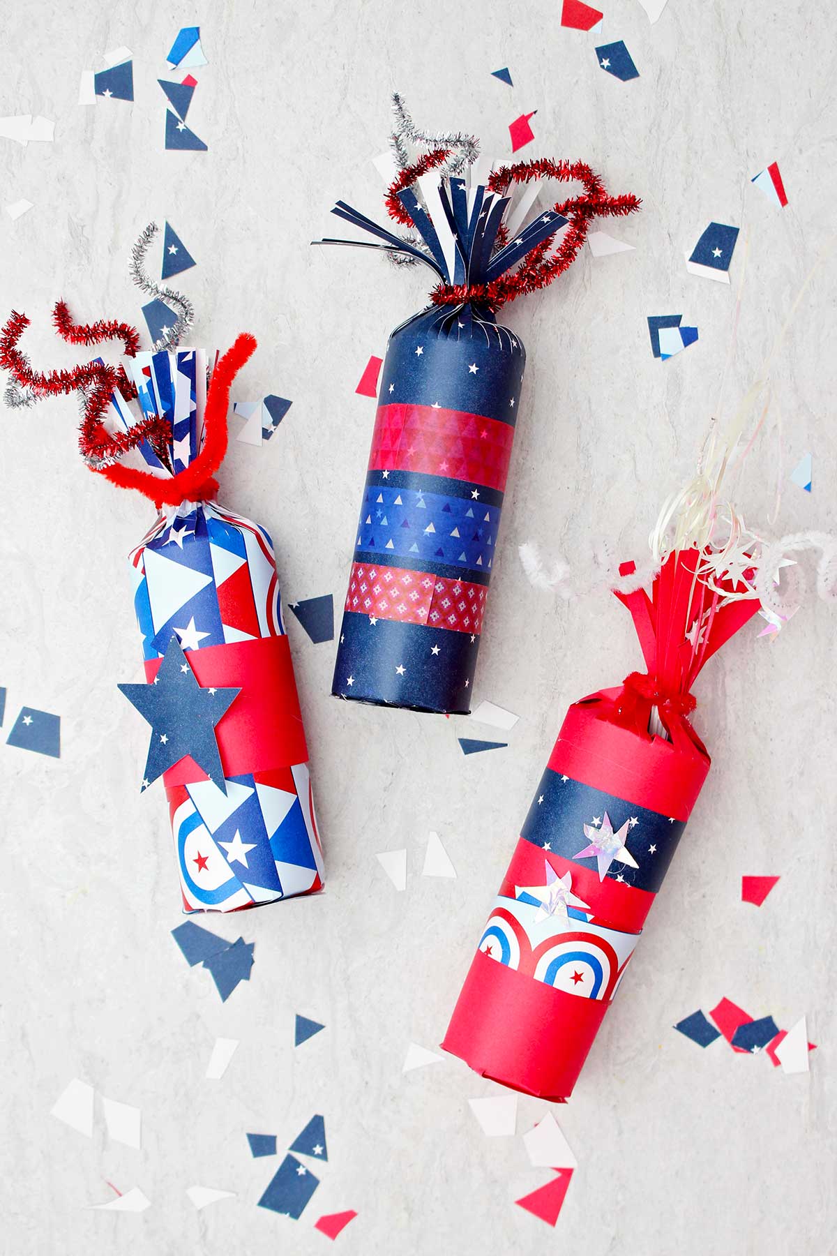 Three 4th of July firecracker treat boxes decorated with red white and blue paper and washi tape with some scraps of festive paper scattered around.