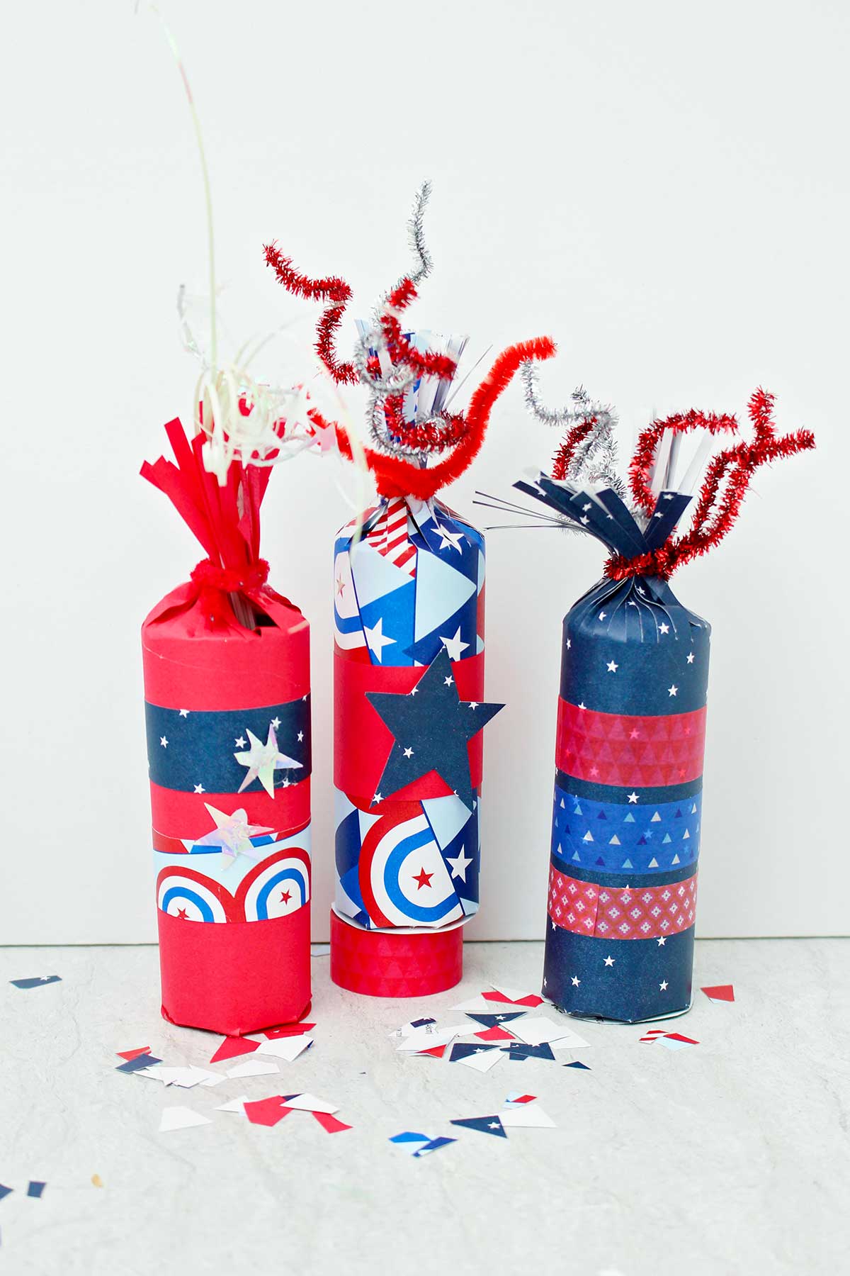 Direct shot of three 4th of July firecracker treat boxes decorated with red white and blue paper and washi tape with some scraps of festive paper scattered around.