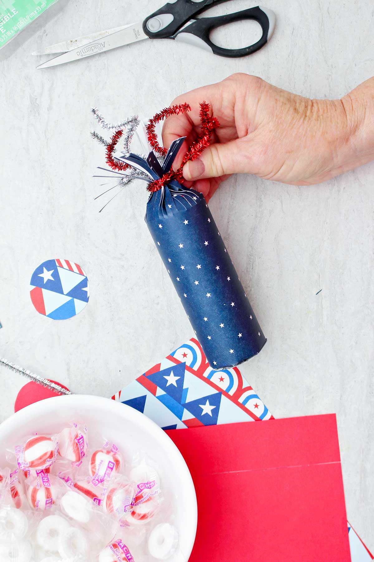 Person wrapping a sparkly red pipe cleaner on top of the blue star patterned firecracker treat box.
