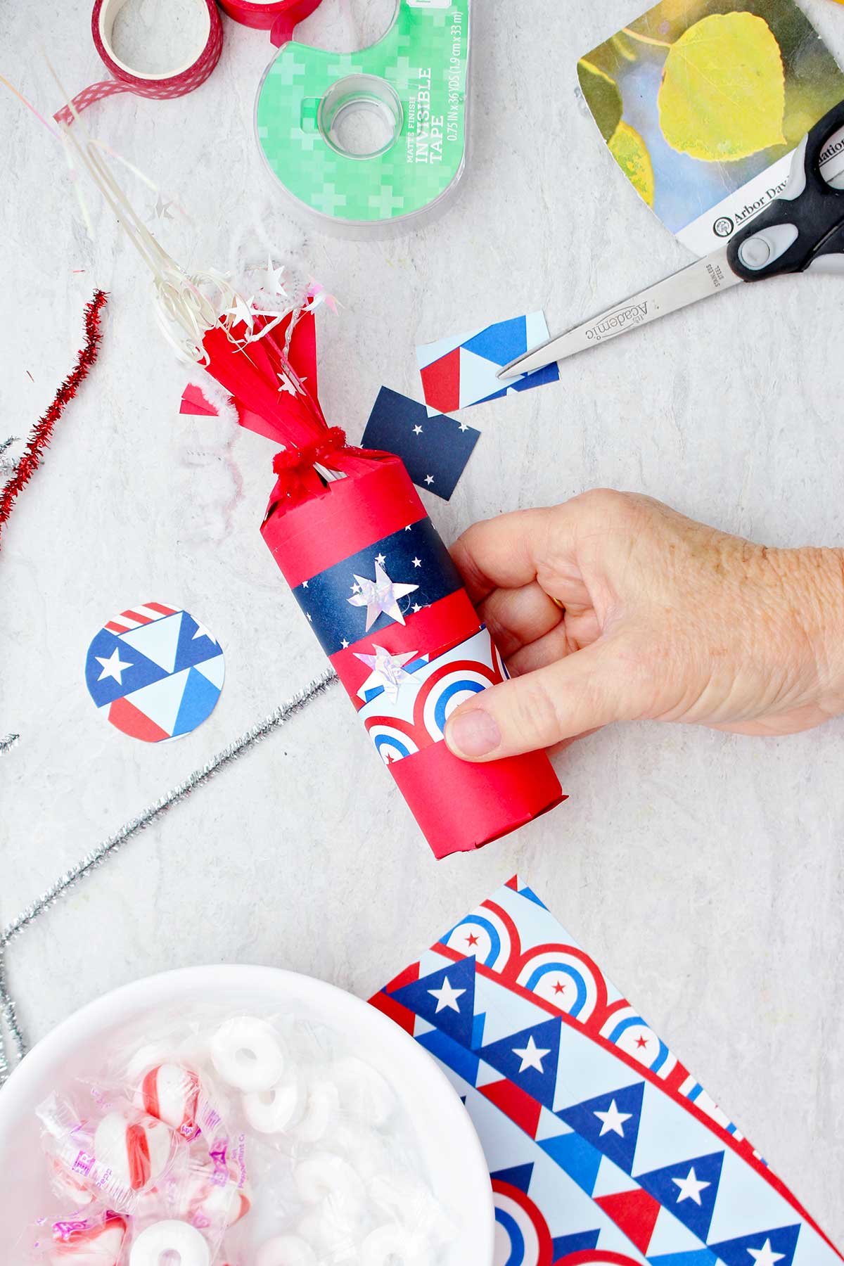 Person decorating red firecracker treat box with patterned scrapbook paper.