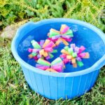Four multi colored sponge bombs in a blue bucket full of water outside.