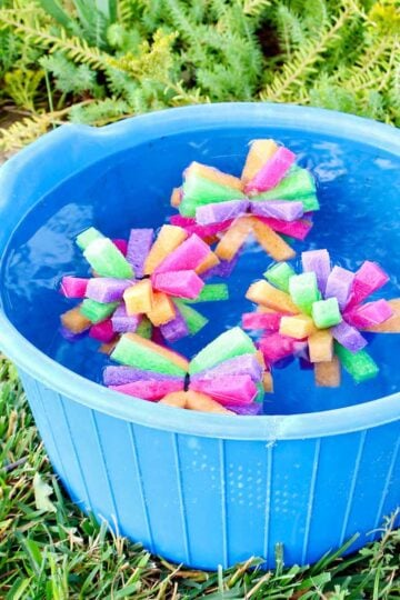 Four multi colored sponge bombs in a blue bucket full of water outside.