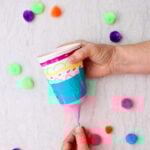 Person pulling the tied end of the balloon on a marshmallow shooter with other pom poms near by.