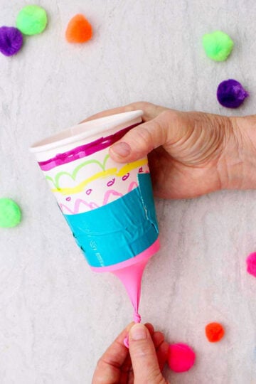 Person pulling the tied end of the balloon on a marshmallow shooter with other pom poms near by.