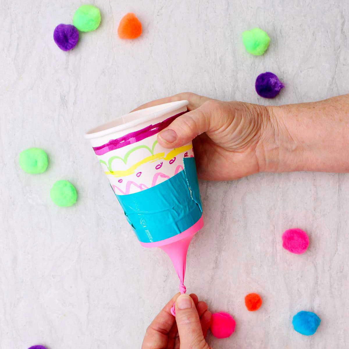 Person pulling the tied end of the balloon on a marshmallow shooter with other pom poms near by.