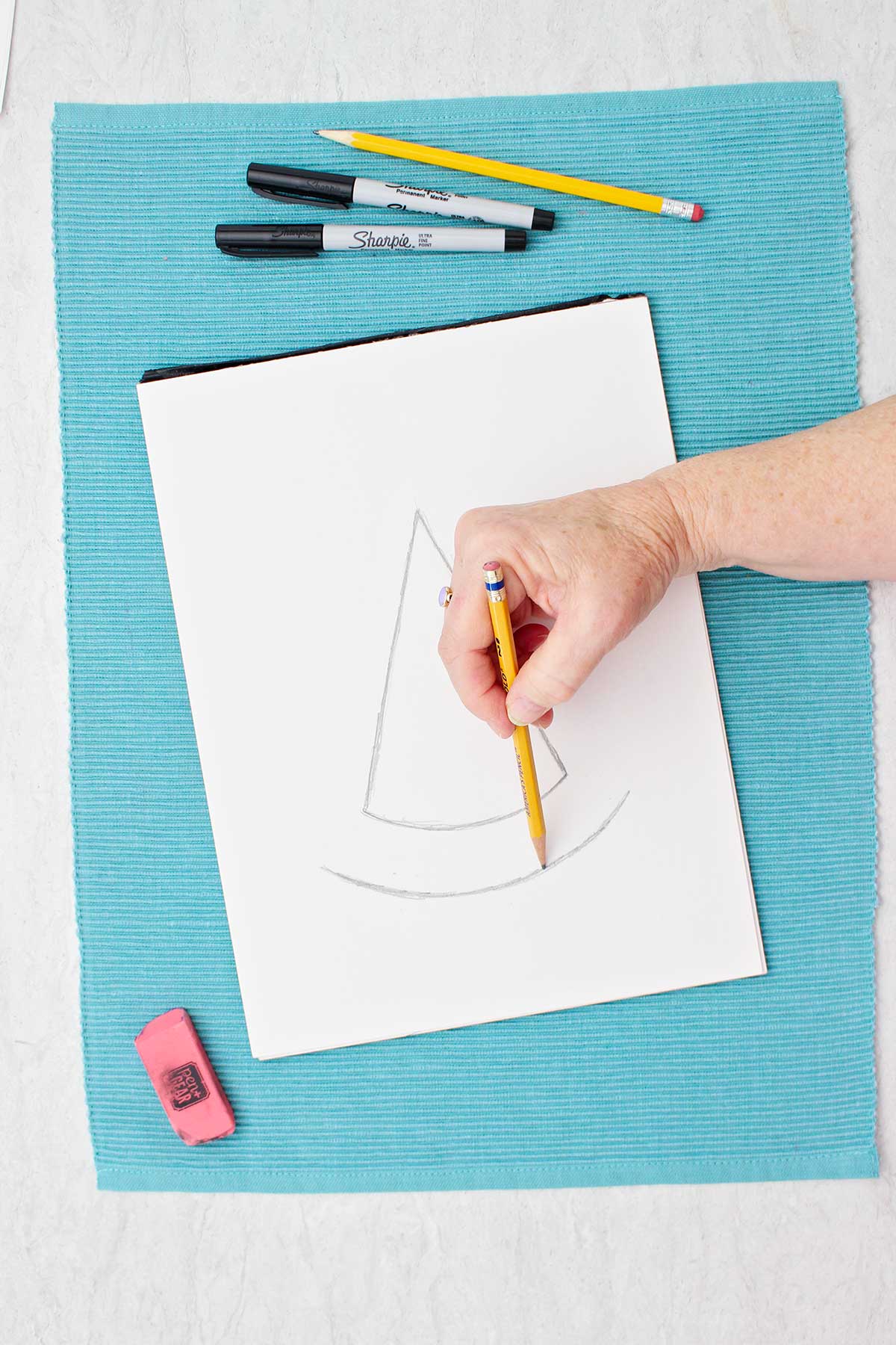 Person sketching bottom curve of witches hat with sketch pad resting on a blue placemat.