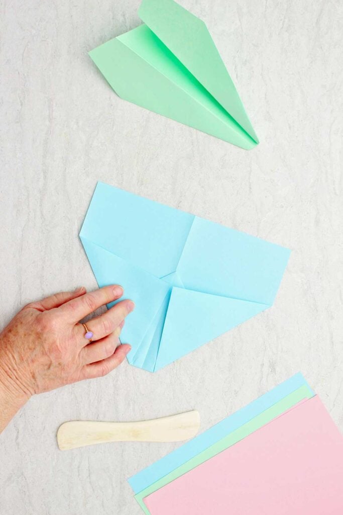 Person folding flaps of a blue paper airplane with green plane, bone folder and paper near by.