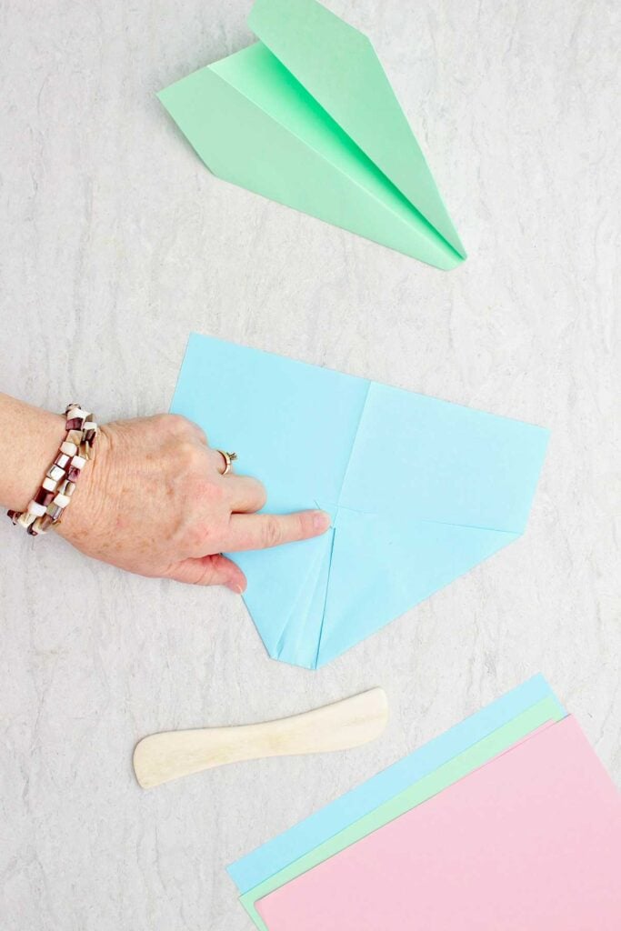 Person folding flaps of a blue paper airplane with green plane, bone folder and paper near by.