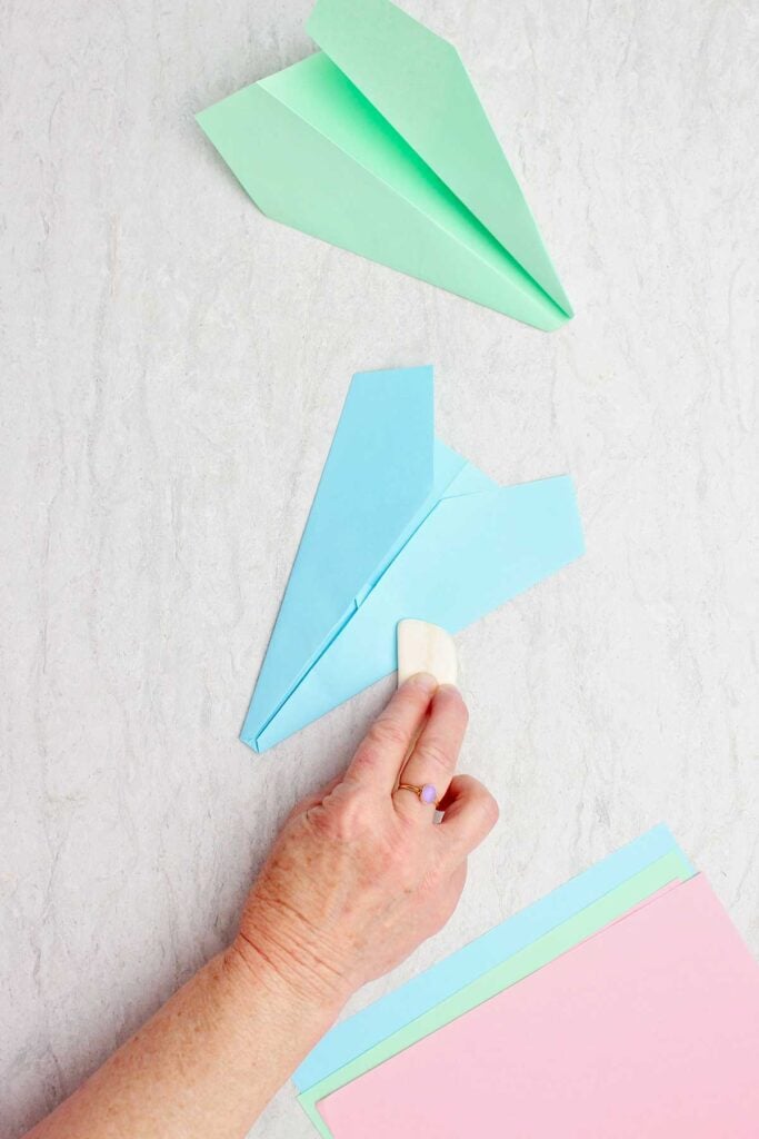 Person using a wooden bone folder to sharpen folds in a blue paper airplane.
