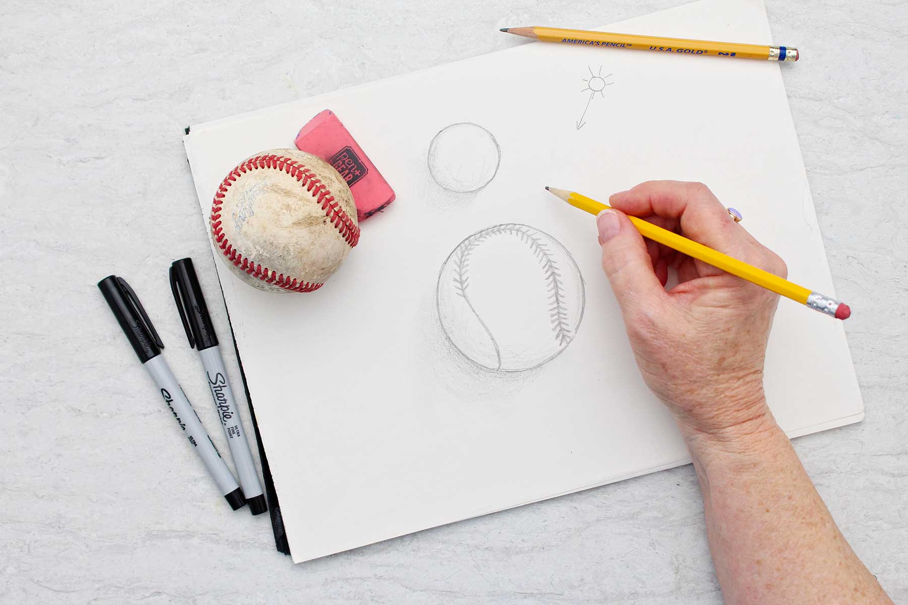 Person lightly sketching stitches in baseball drawing with other drawing supplies and baseball near by.