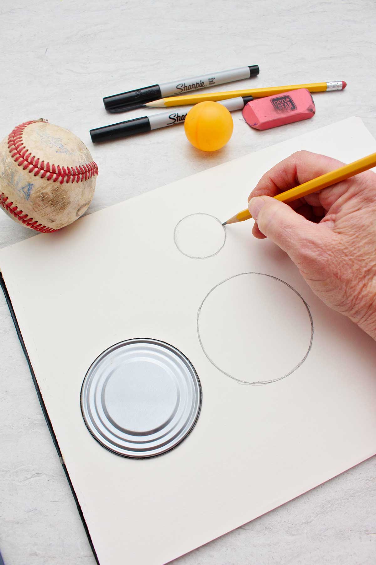 Person drawing a small circle near a larger circle with a pencil with a tin can lid, pencil, eraser, sharpies, ping pong ball and baseball near by.