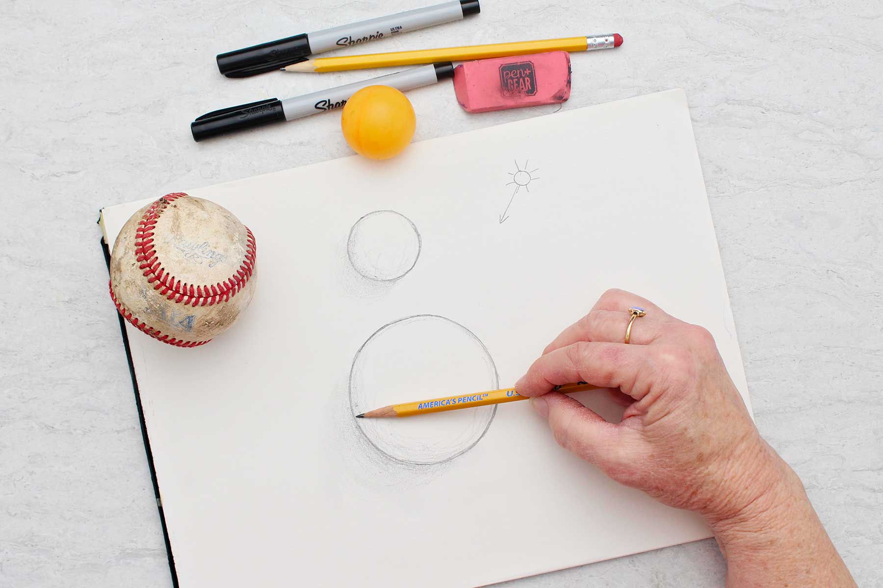 Overhead view of person shading left side of sphere sketched with a pencil.