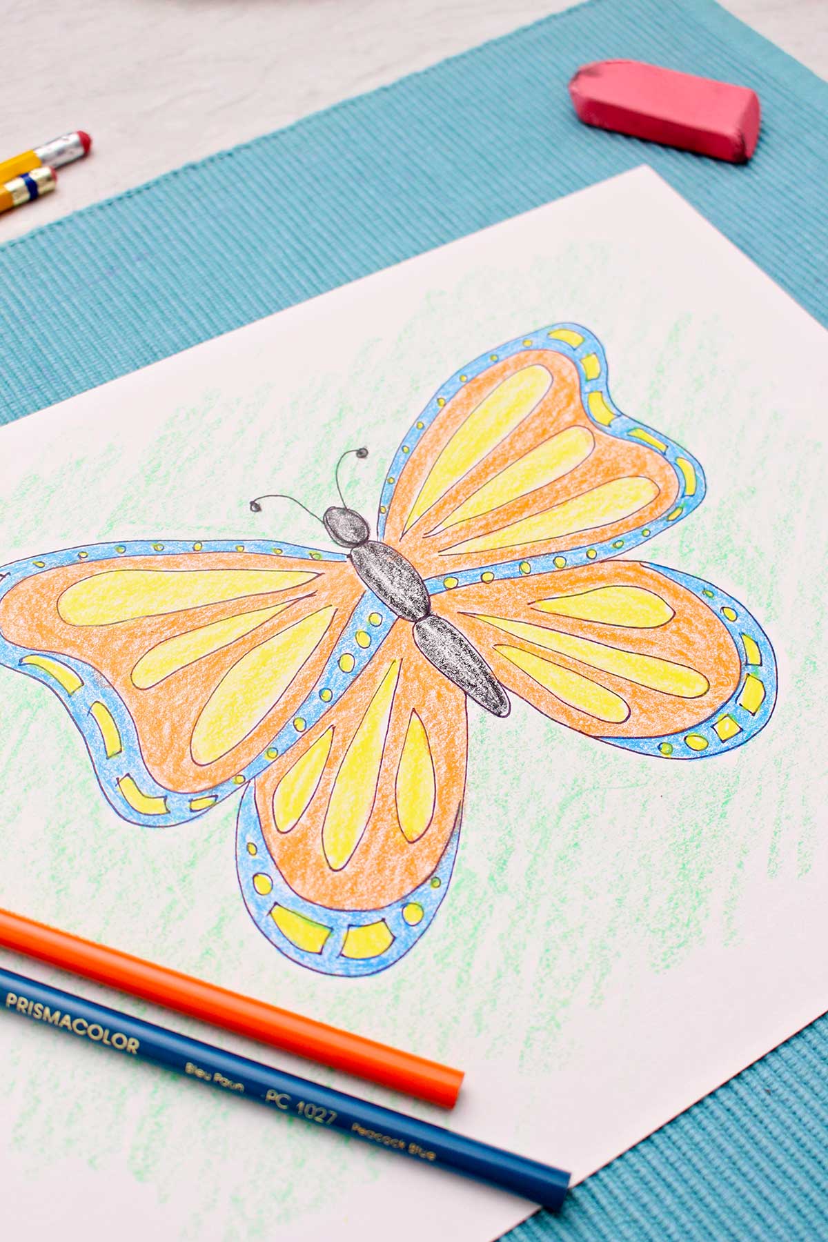 Blue, yellow and orange colored butterfly drawing on blue placemat with colored pencils and sharpies around.