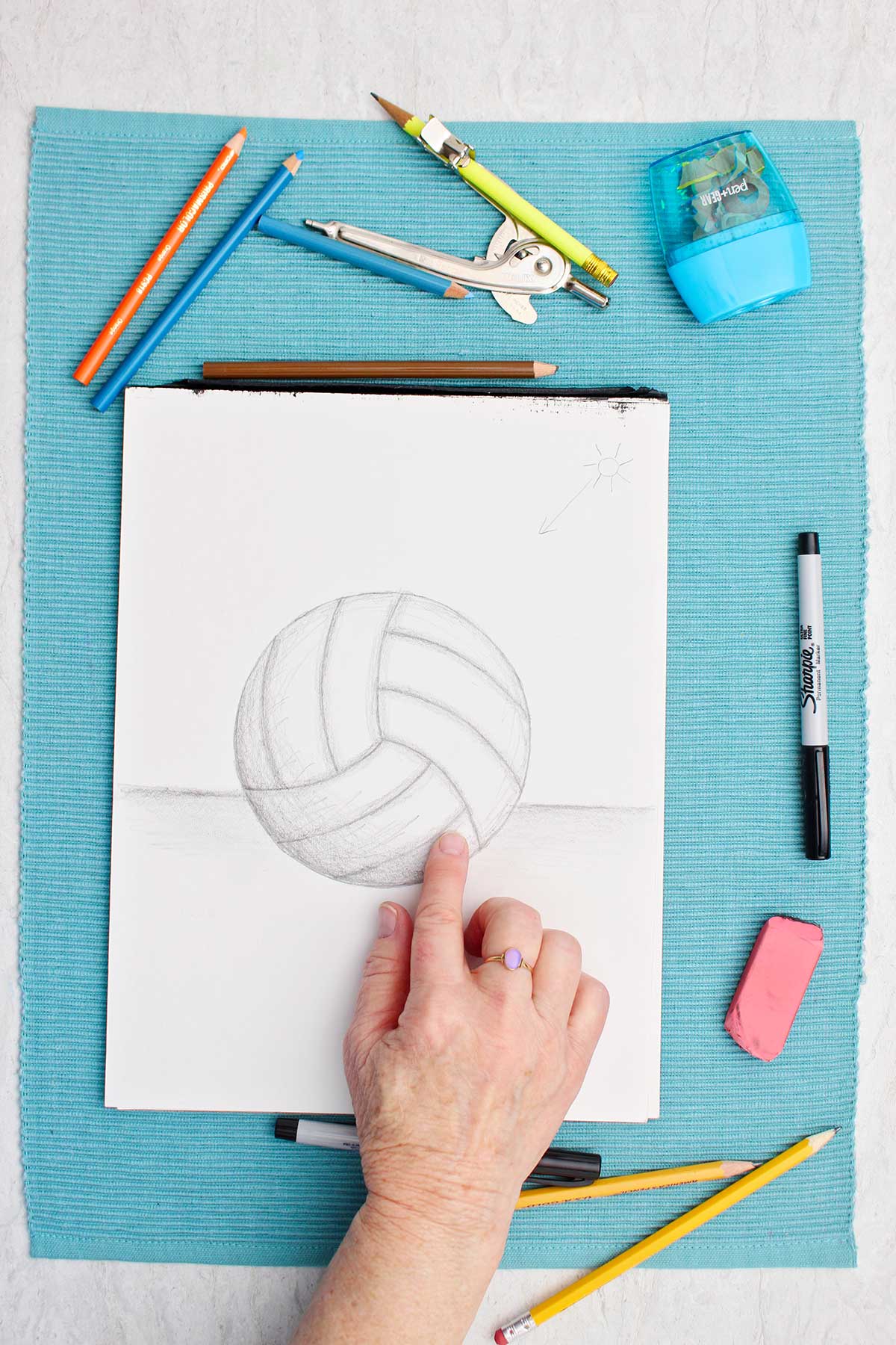 A drawing of a volleyball in pencil with a person shading with finger with supplies near by on a blue placemat.