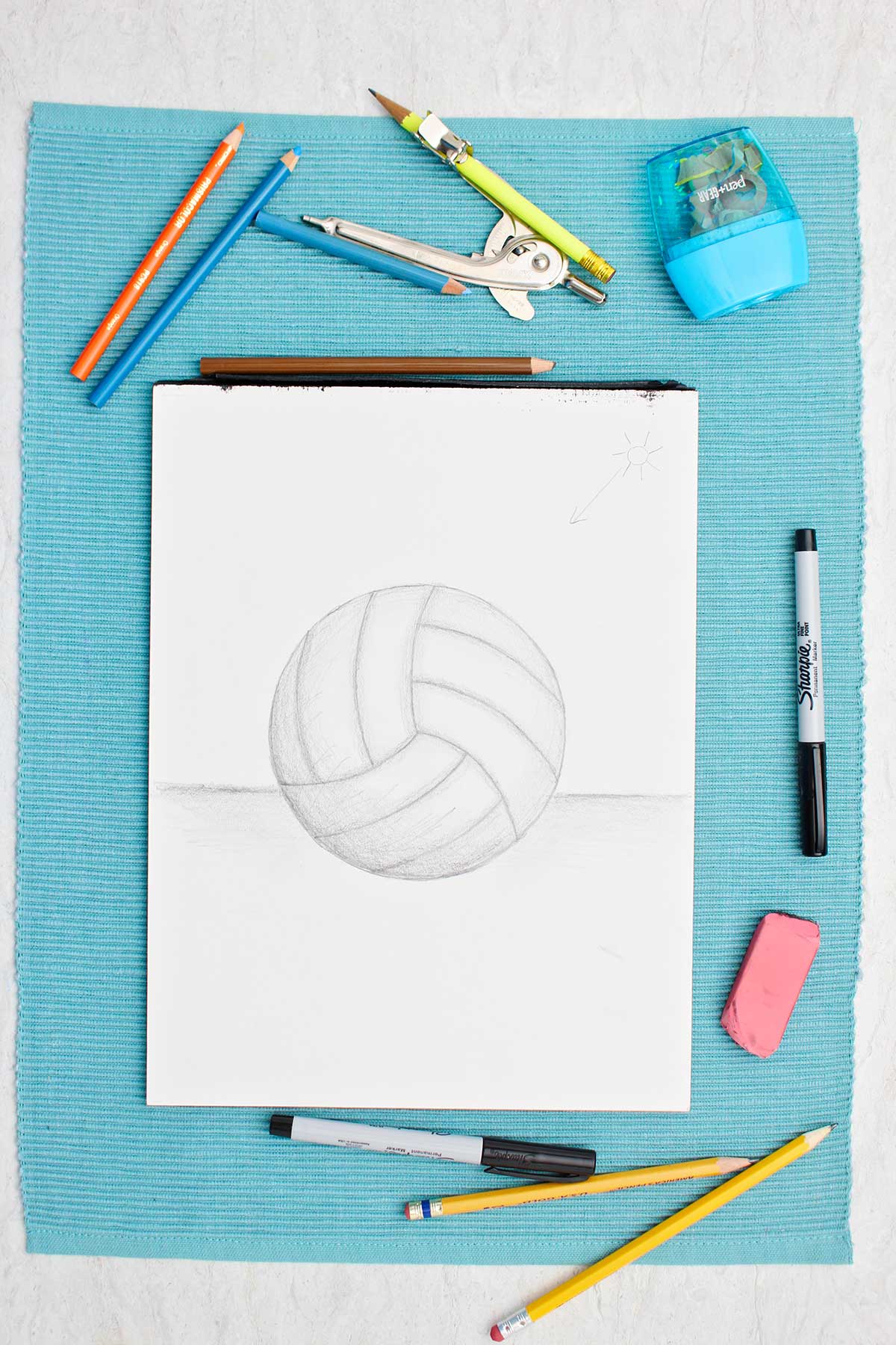 A drawing of a volleyball in pencil with shading with supplies near by on a blue placemat.