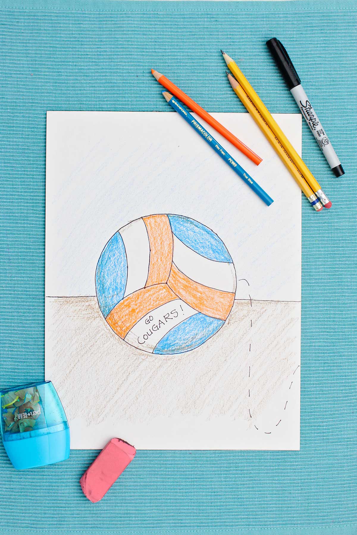 Drawing of volleyball colored in with blue and orange colored pencils with supplies near by on a blue placemat.