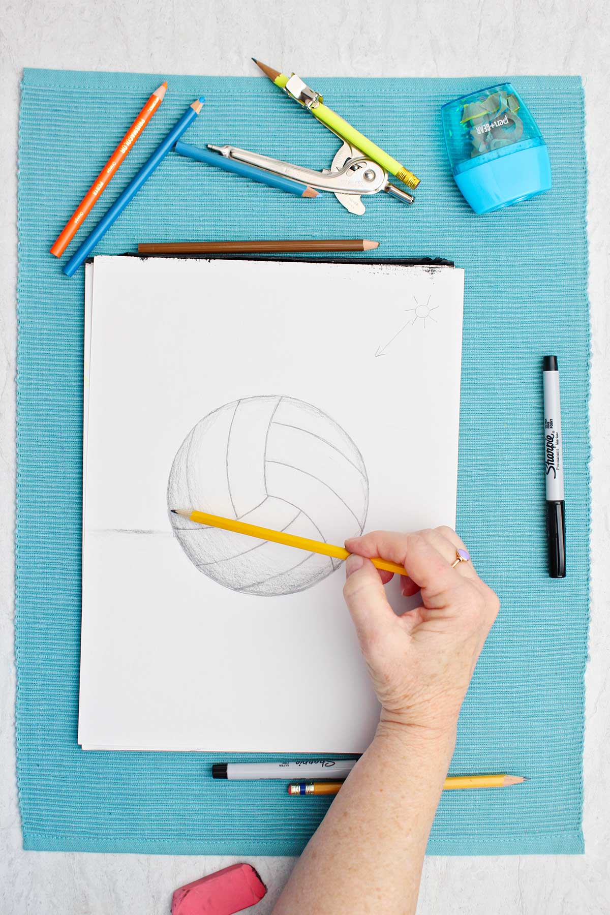 A drawing of a volleyball in pencil with a person shading with finger with supplies near by on a blue placemat.