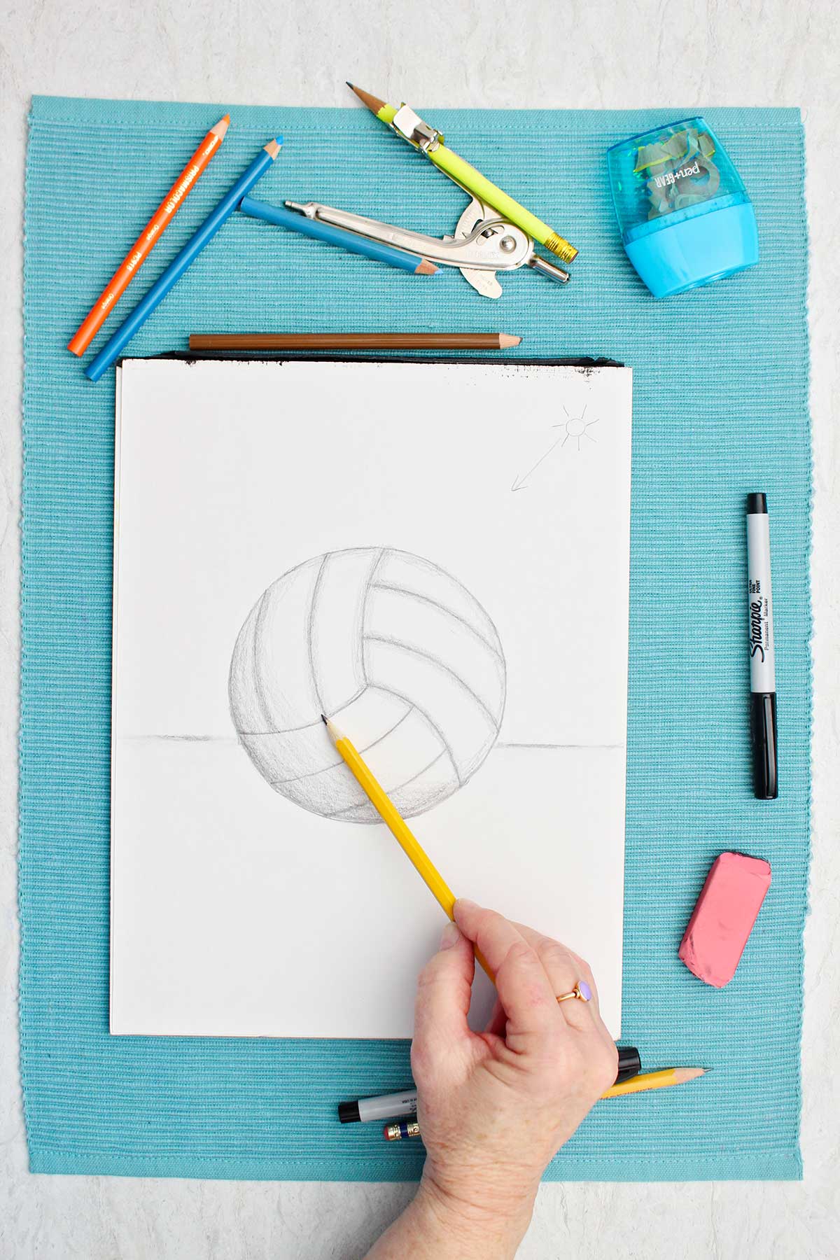 A drawing of a volleyball in pencil with a person shading with pencil with supplies near by on a blue placemat.