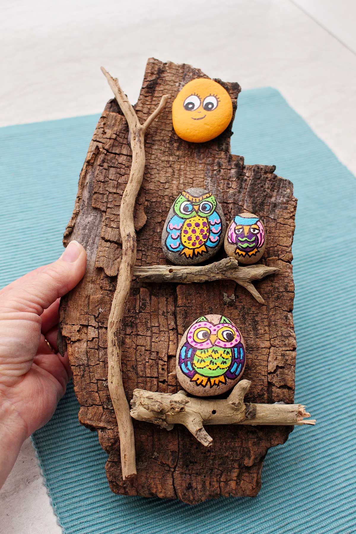 Person holding three painted rocks of colorful owls and an orange painted rock attached to a piece of wood.