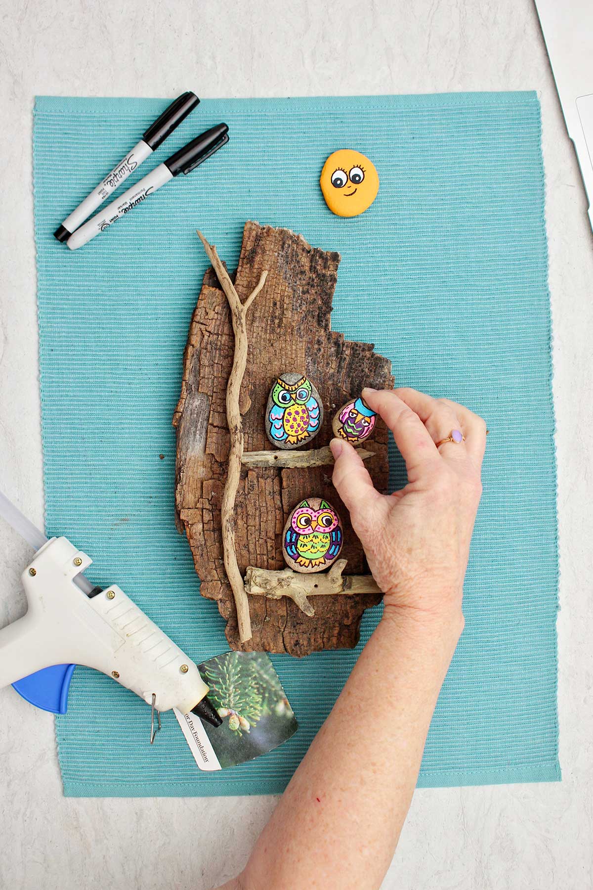 Person gluing down smallest painted owl rock on a branch attached to a piece of wood.