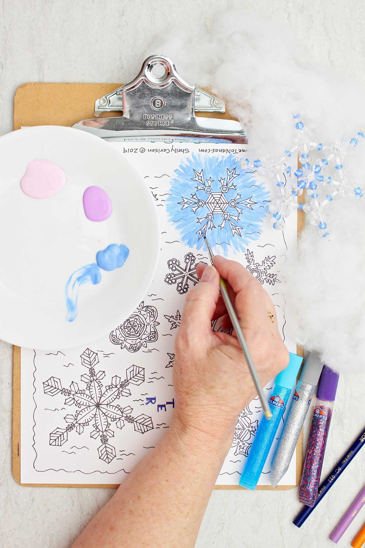 Person using blue paint to paint around the outside of a snowflake on free printable snowflake coloring page.