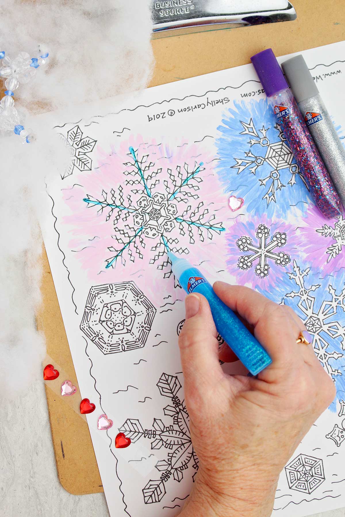 Person using glitter glue to paint the details of a snowflake on free printable snowflake coloring page.