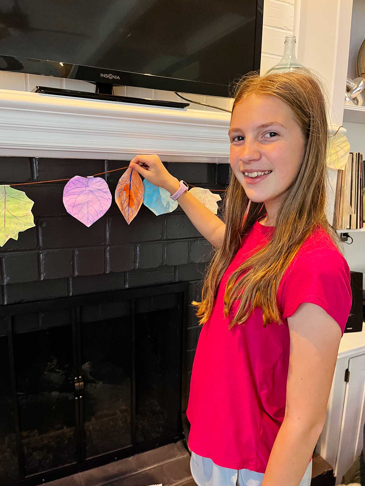 A young girl with brown hair and a pink shirt hangs leaf rubbing garland on a mantle.
