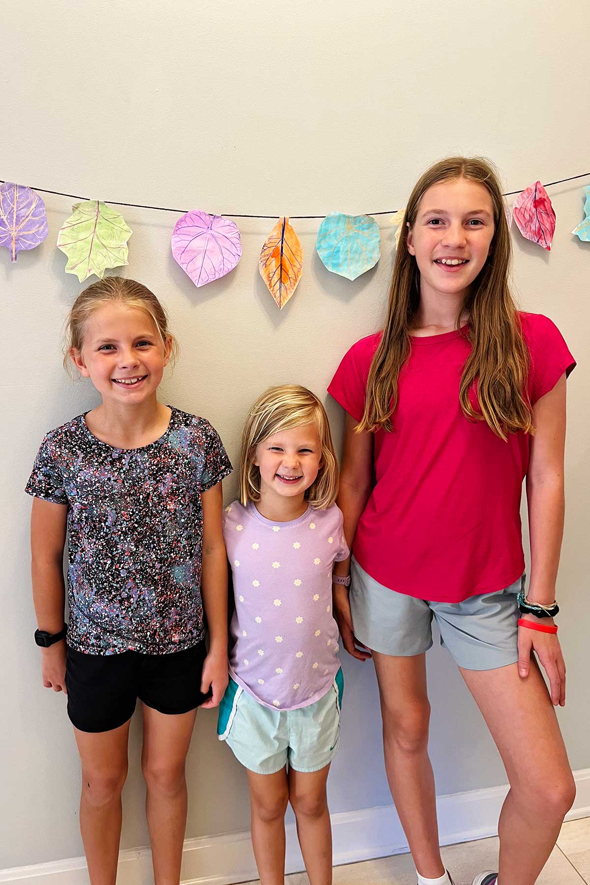 Three young girls smile stand in front of their leaf rubbing garland.