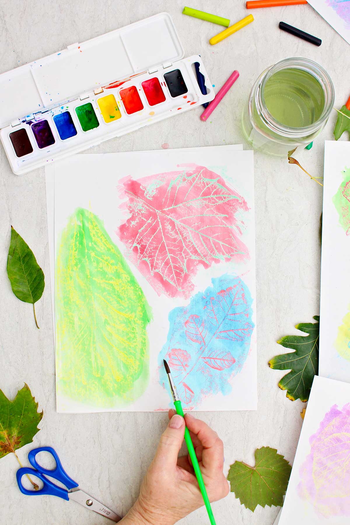 A person watercolors over leaf rubbings with scattered leaves and watercolors near by.