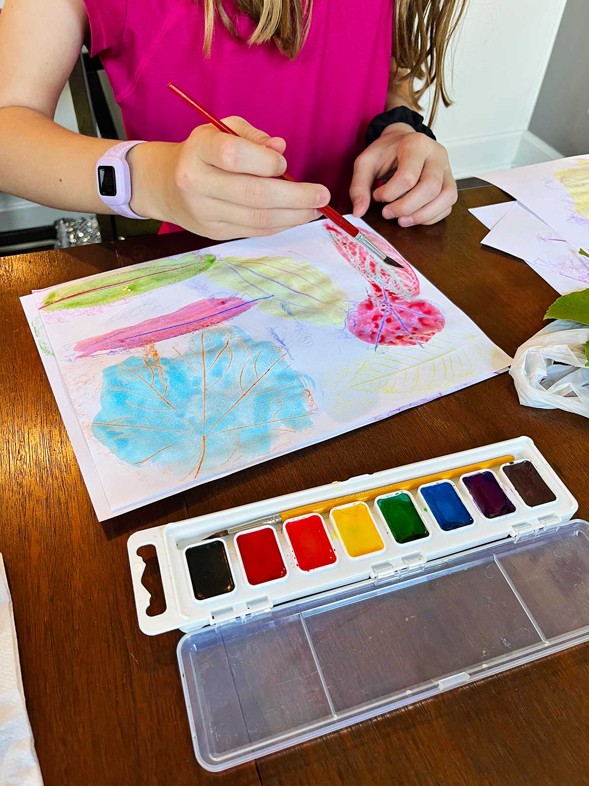 A young girl with a pink shirt watercolors over leaf rubbings at a table.
