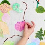A person securing a red and purple leaf rubbing on a piece of yarn with tape with other leaf rubbings scattered around.