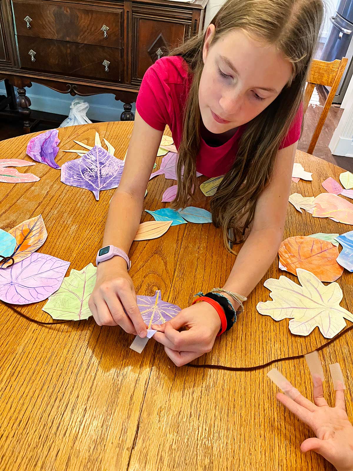 A young girl with brown hair and a pink shirt tapes leaf rubbings to a piece of brown yarn.