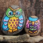Close up of two colorfully painted owl rocks on a branch attached to a piece of wood.