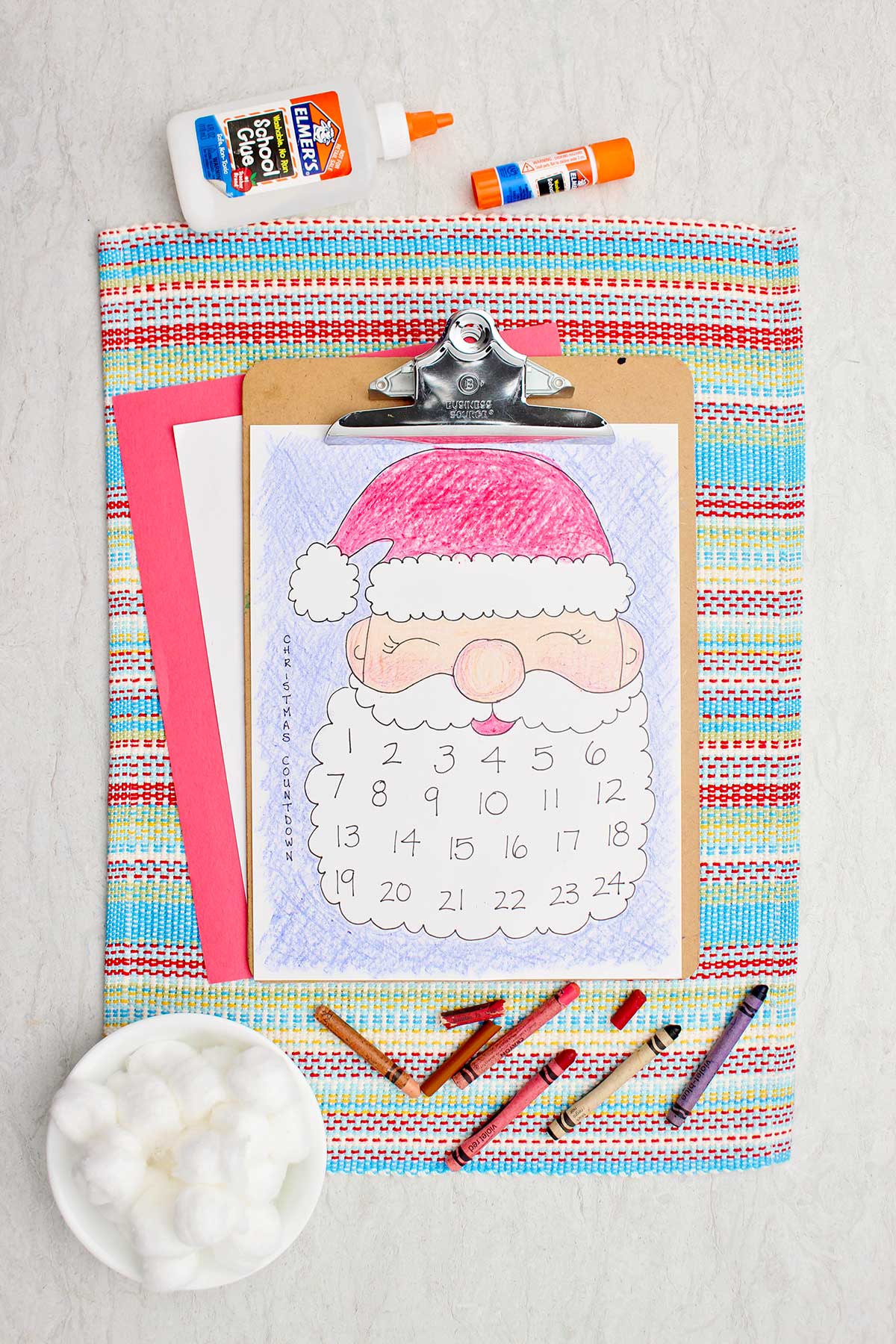 A colored Santa calendar printout on a clip board resting on a placemat with glue, crayons and cotton balls near by.