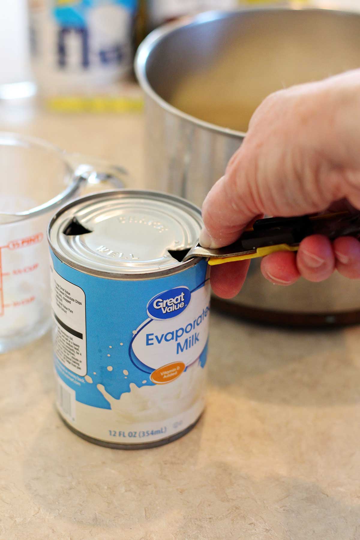 A person cutting open a can of evaporated milk with a saucepan in the background.