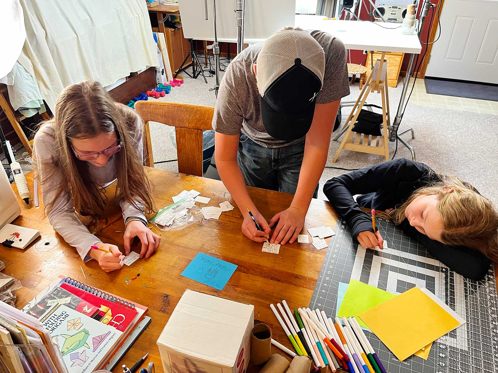 A boy and two girls writing Would You Rather questions on small pieces of paper.