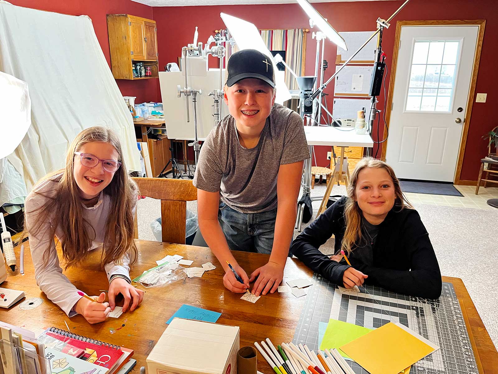 A smiling boy and two girls writing Would You Rather questions on small pieces of paper.
