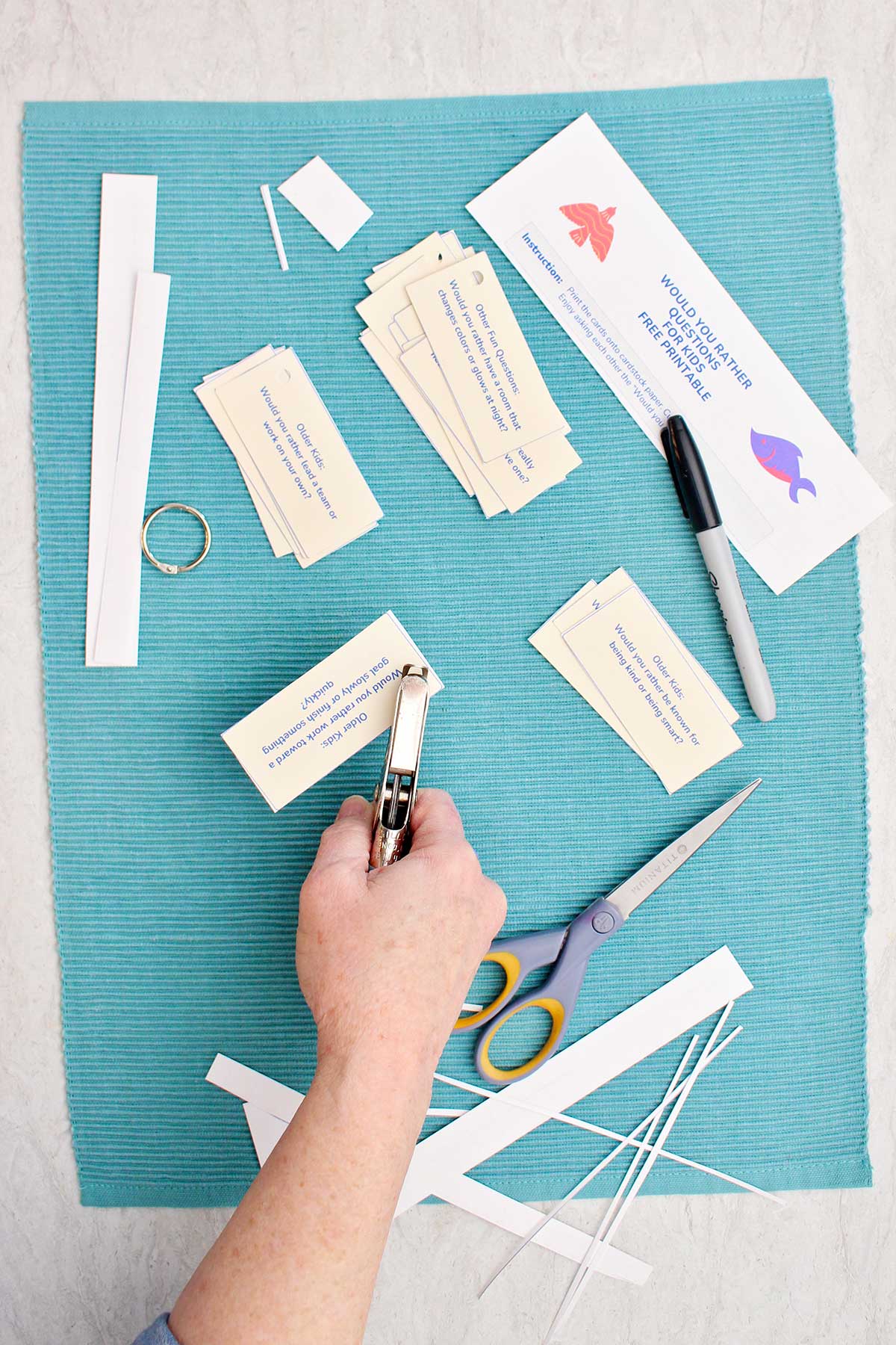 Someone punching a hold in a Would You Rather question on a small notecard, with cut pieces of paper, a pair of scissors, a hole punch, and a marker nearby.