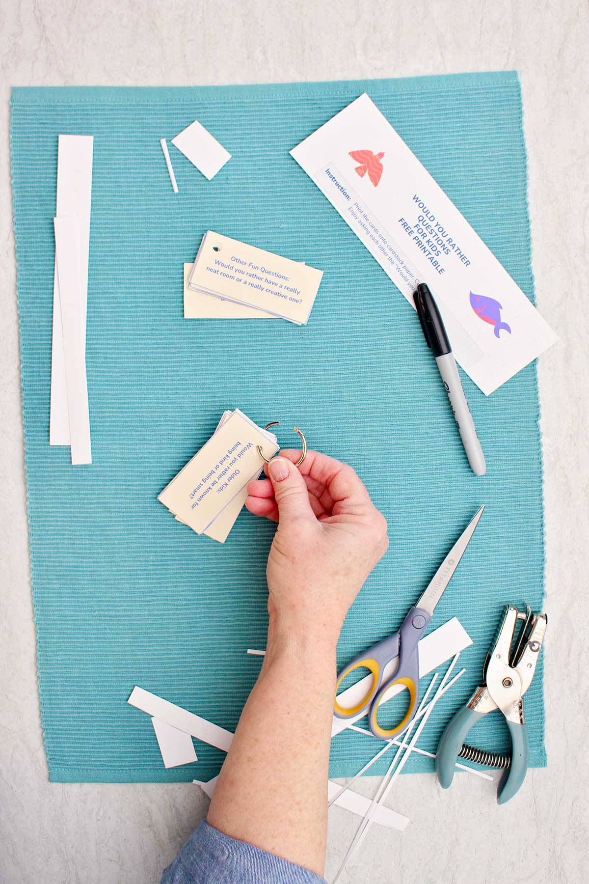 Someone closing the ring on a stack of Would You Rather questions with cut pieces of paper, a pair of scissors, a hole punch, and a marker nearby.