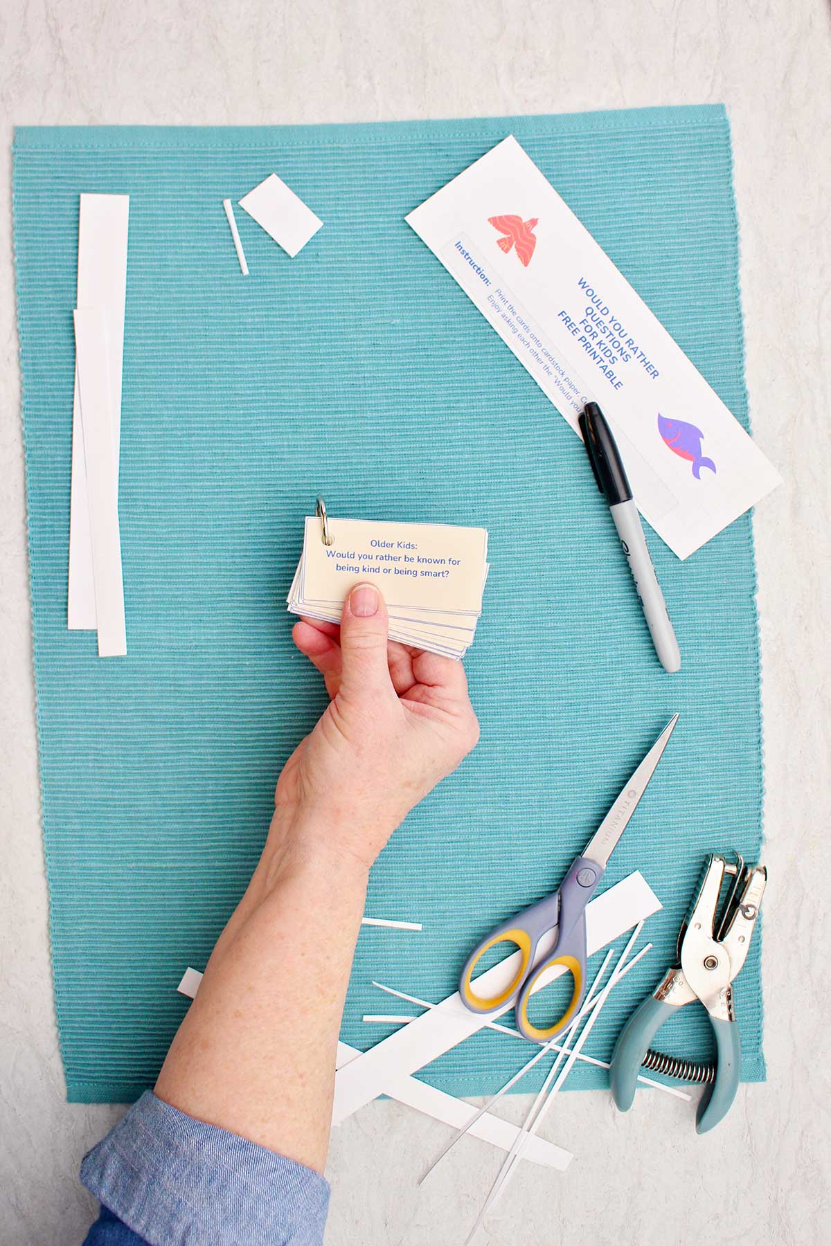 Someone holding a stack of Would You Rather questions with cut pieces of paper, a pair of scissors, a hole punch, and a marker nearby.