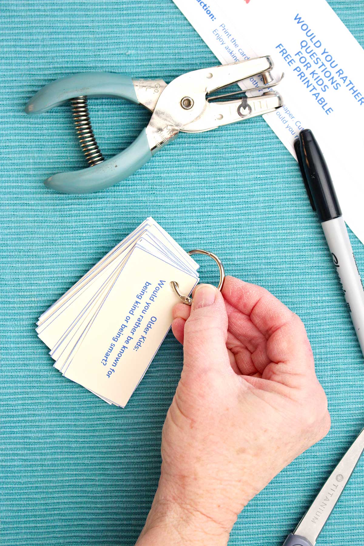 A person closing a ring on a stack of Would You Rather Questions next to a marker and a hole punch.