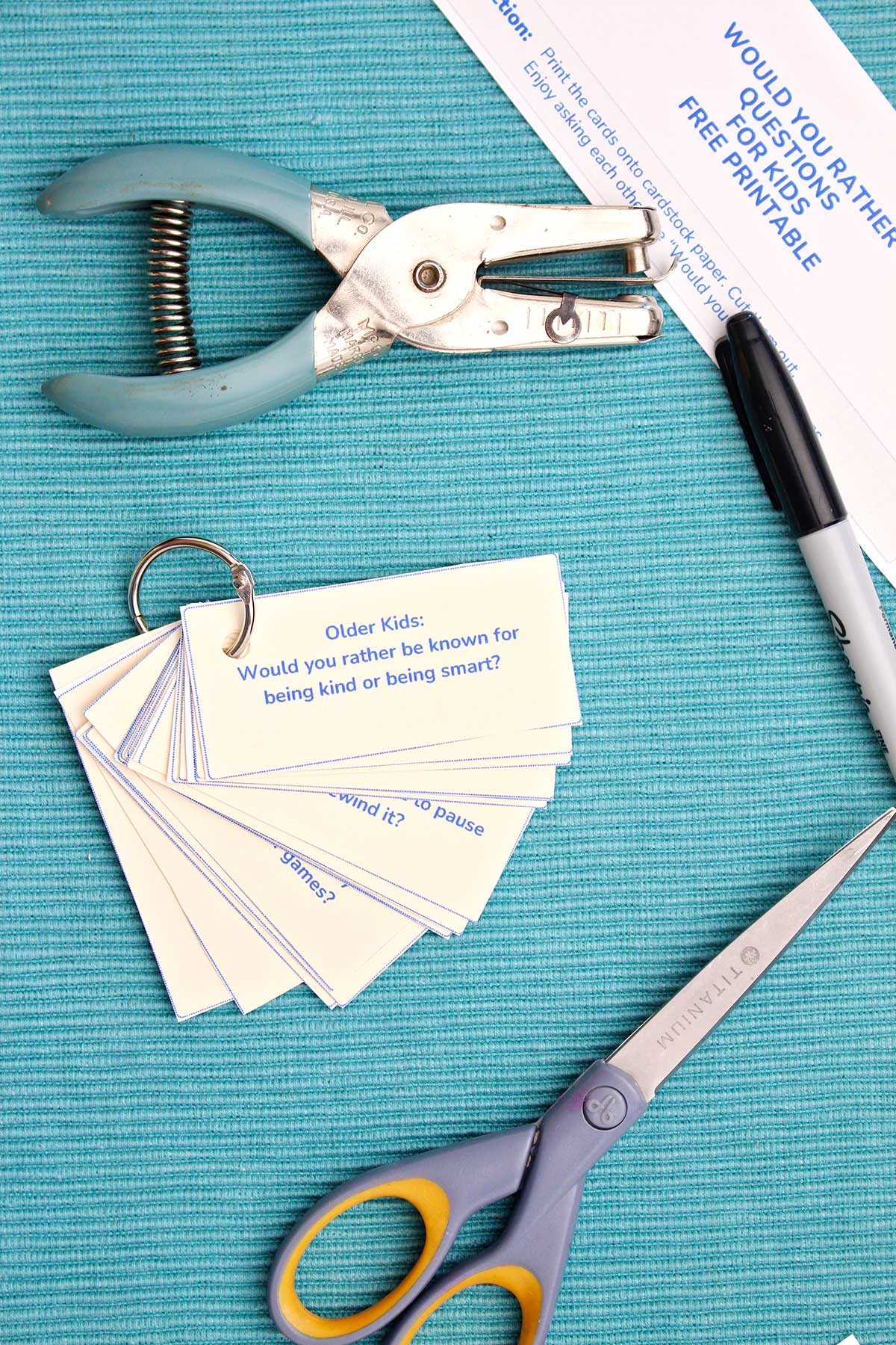 A ring of Would You Rather Questions next to a marker, pair of scissors, and a hole punch.
