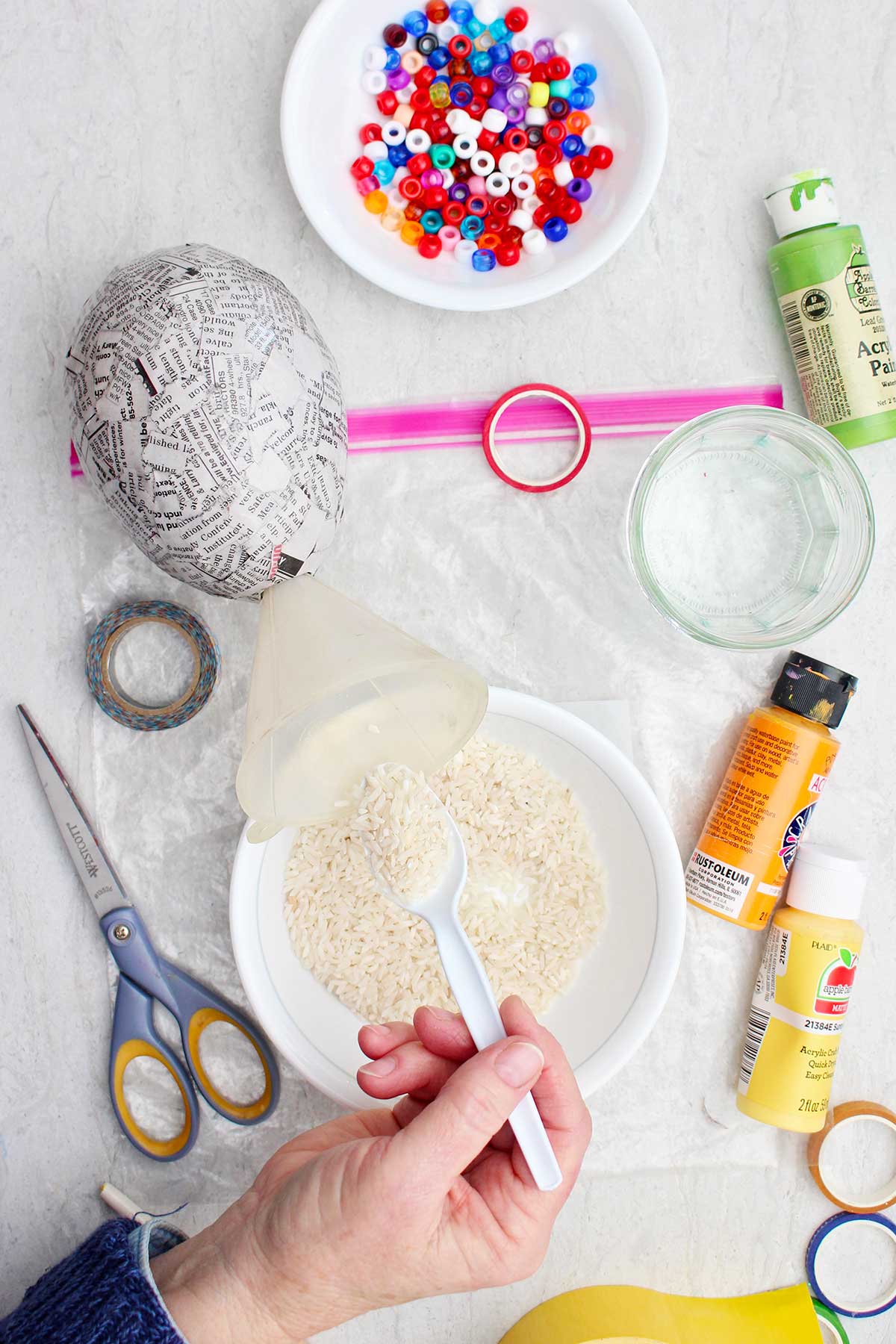 Person filling their news paper maraca with rice through a funnel with other supplies near by.