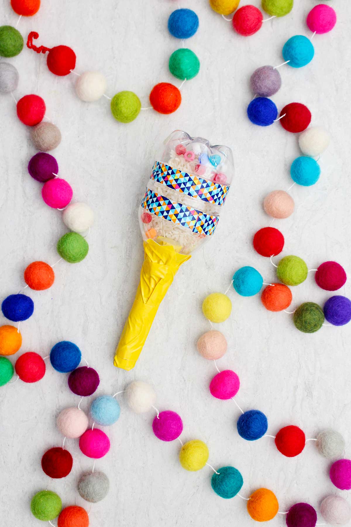 Maraca made with a small water bottle filled with rice and beads with yellow handle with colorful pom poms near by.