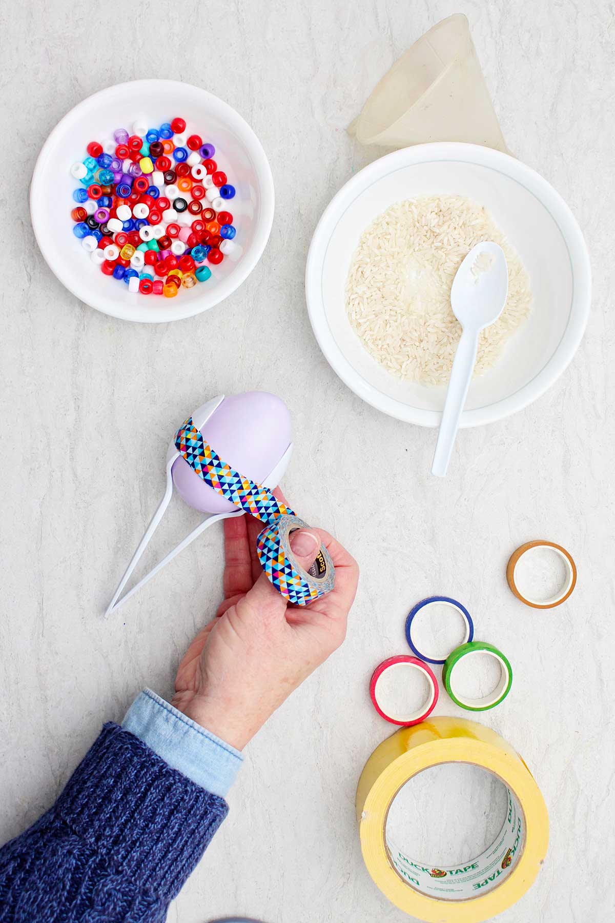 Person securing two plastic spoons around a plastic egg with colorful washi tape.